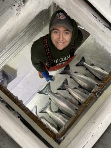 A woman stands in a freezer looking up at the camera, with fish spread before her on a table.