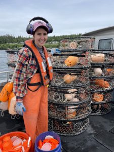Emily Buckner wearing orange waders, a life vest, and headphones stands near a stack of crab pots.