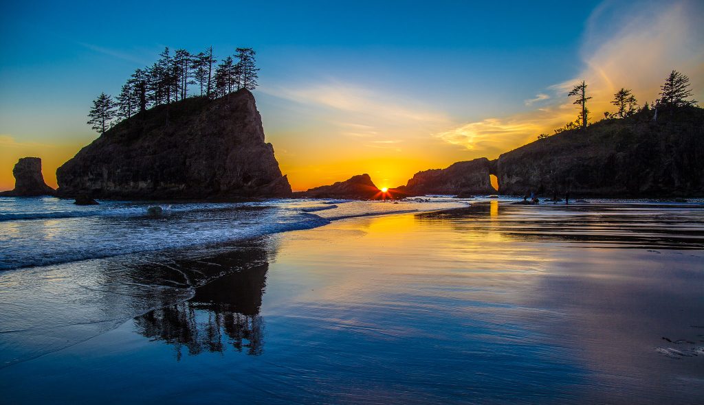 Sunset on the beach with large rock formations covered with trees in the background.