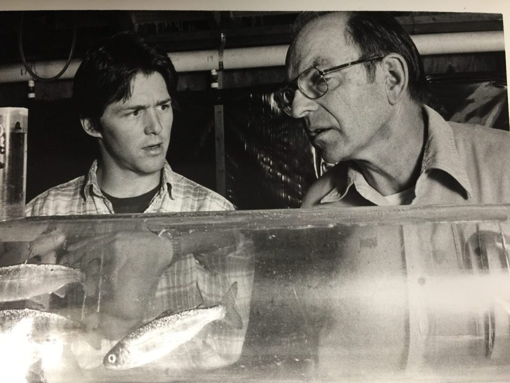 A student listens to a researcher, the two of them standing before fish in a tank.