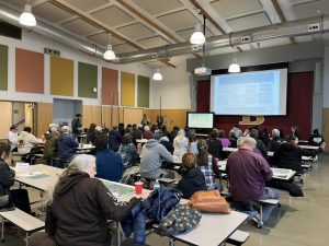 A crowd of community members sits at tables facing a projector screen.