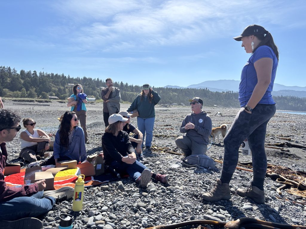 Vanessa Castle stands on the right, addressing a group of fellows seated on a rocky beach