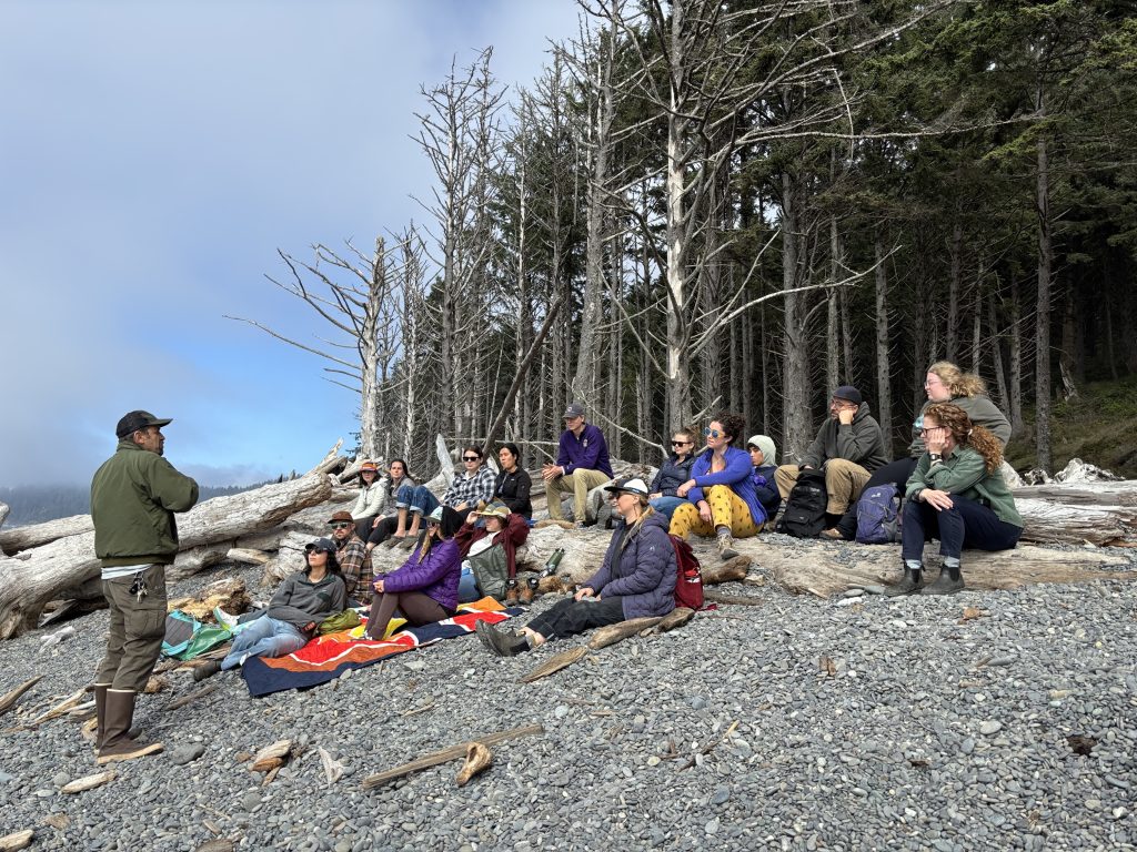 Dr. Steve Fradkin, standing, addresses a group of seated fellows on rocky beach, with forest beyond.
