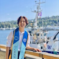 Isaac Olson stands in a graduate sash in front of a UW research vessel