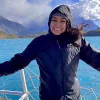 Mariah Ricci on a boat with blue water, mountains, and clouds behind.
