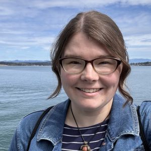 WSG Science Communications Fellow Emily Bjornsgard smiles at the camera, wearing glasses and a denim jacket in front of the Puget Sound.