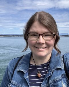 WSG Science Communications Fellow Emily Bjornsgard smiles at the camera, wearing glasses and a denim jacket in front of the Puget Sound.