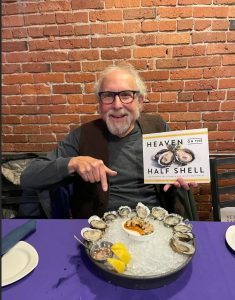 David Gordon holds a copy of Heaven on the Half Shell while sitting in front of a plate of oysters on the half shell